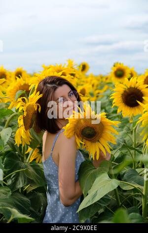 Beautiful young woman in a rural field scene outdoors standing between sunflowers, enjoying sunset at summer evening Stock Photo