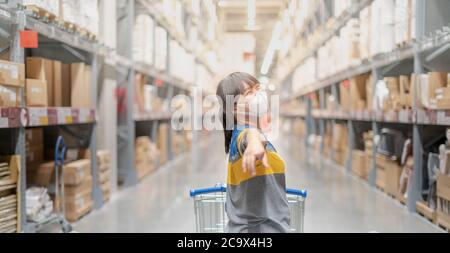 The asian girl wearing surgical mask pushing the trolley cart shopping the decorate funiture for interior inside the house in the big warehouse store. Stock Photo