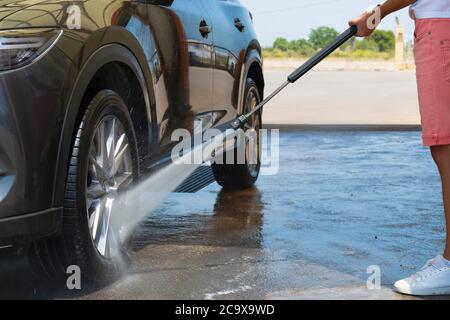 Close up of a man holding a high pressure water spray wand washing the front wheel of a car on an out of focus background. Car washing and cleaning co Stock Photo