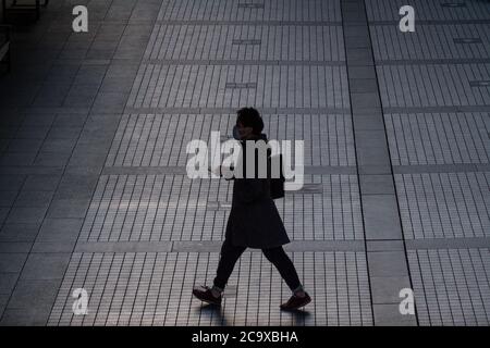 A man in silhouette and using a smartphone and wearing a face-mask walks across a plaza in Shinjuku, Tokyo, Japan Stock Photo