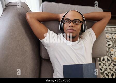 Portrait of smiling dreamy young Black man resting on sofa in headphones and looking away Stock Photo
