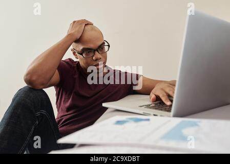Pensive young African-American businessman in casual clothes analyzing financial report on laptop screen Stock Photo