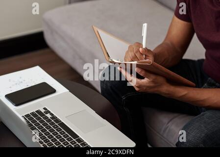 Close-up image of university student watching online webinar on laptop and taking notes in copybook Stock Photo