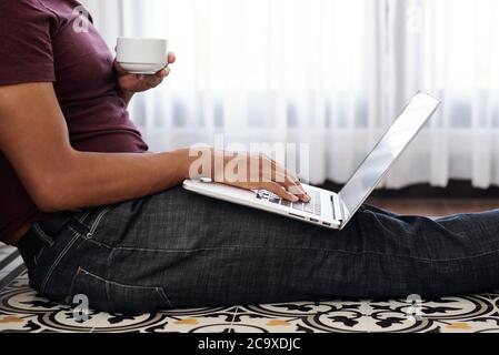 Cropped image of young man sitting on the floor at home, drinking morning coffee and answering e-mails Stock Photo