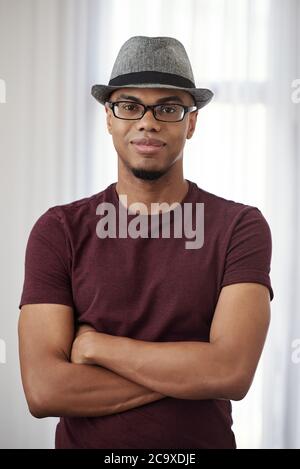 Portrait of handsome smiling young Black man in fedora hat and glasses crossing hands and smiling at camera Stock Photo