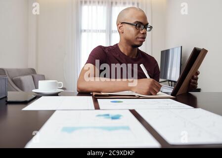 Serious young African-American businessman analyzing report on tablet computer and taking notes in planner Stock Photo