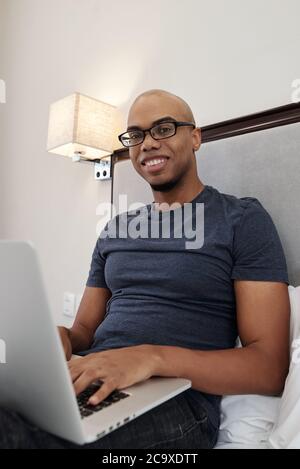Portrait of positive young Black man sitting on bed, working on laptop and smiling at camera Stock Photo