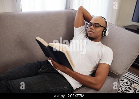 Smiling young African-American man in headphones relaxing on sofa and reading interesting novel Stock Photo