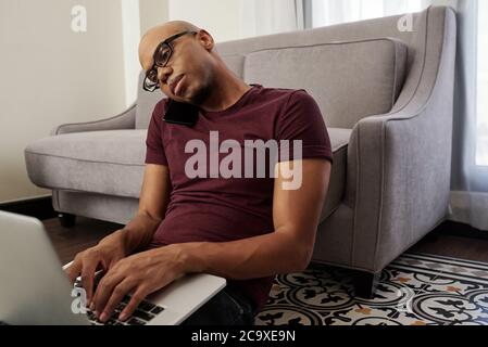 Serious young Black programmer sitting on the floor, talking on phone with client and working on laptop Stock Photo