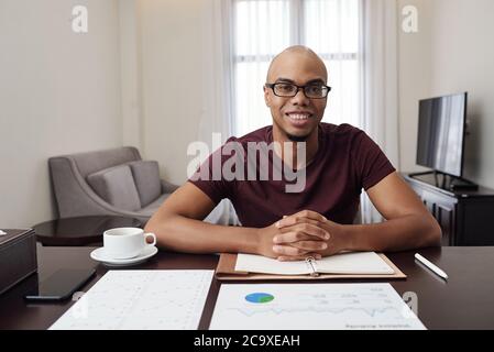 Potrait of handsome smiling young Black businessman in glasses sitting at desk at home and working with financial documents Stock Photo