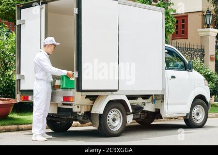 Delivery man in white uniform taking plastic crate with glass milk bottles out of van trunk Stock Photo