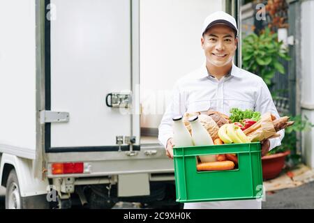 Portrait of positive young Vietnamese courier holding plastic box with fresh fruits, bread and bottled milk Stock Photo