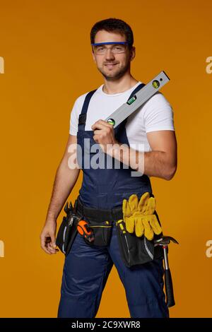 Young smiling caucasian repairman worker in safety glasses and uniform holding builder level. handyman wearing tool belt isolated on orange studio background Stock Photo