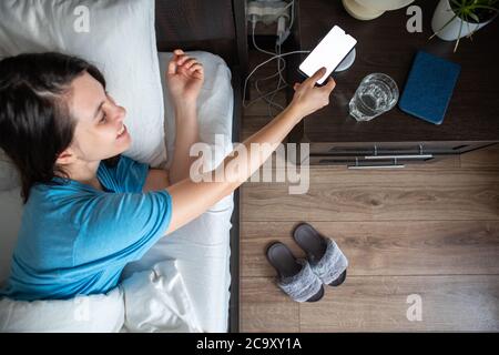 awake woman taking phone from wireless charger from bedside table Stock Photo