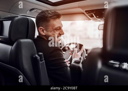 Back view of happy businessman driving automobile during sunny day and using smartphone. Rent and trade-in concept Stock Photo