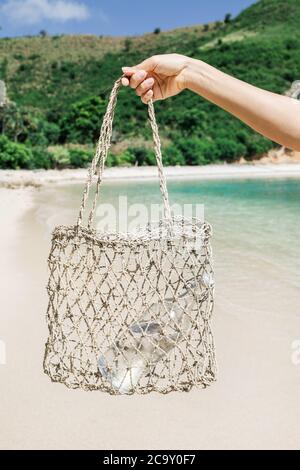 Woman holding straw wicker reusable bag with glass bottle for water. Plastic free and zero waste lifestyle. White sand beach with clear turquoise water on background. Stock Photo