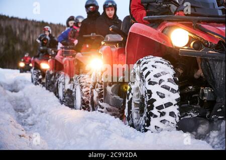 Focus on the wheel. People riding red all-terrain vehicles down snowy hill. Quad riders driving quad bikes with black snowy wheels on snow-covered trail. Concept of winter activities and quad biking. Stock Photo
