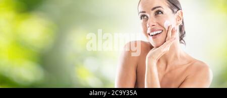 Beautiful young woman applying cream to her face. Skincare spa wellness and cosmetics concept. Blurred green panoramic banner. Stock Photo