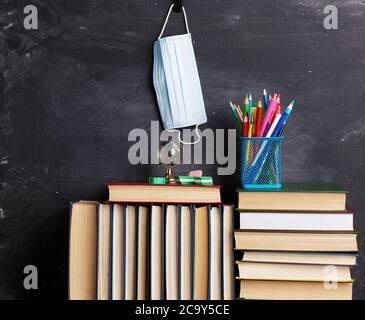 stack of assorted books, stationery and disposable medical mask hanging on black chalk board, back to school Stock Photo