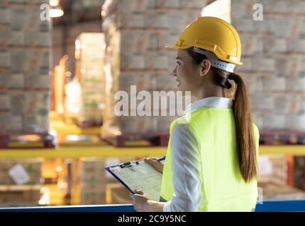 Side view of asian female warehouse worker with archboard flip chart look inventory with dustrubution warehouse in background. Reopening business ware Stock Photo