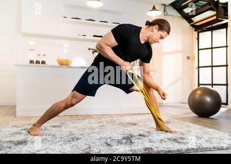 White caucasian fit sporty muscular man doing exercise with stretching band in living room at home for mental health and meditation. Working out fitne Stock Photo