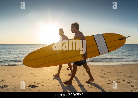 Two mature men walking with surfboards on beautiful beach enjoying paradise and retirement lifestyle. Attractive fit senior adults friends having fun