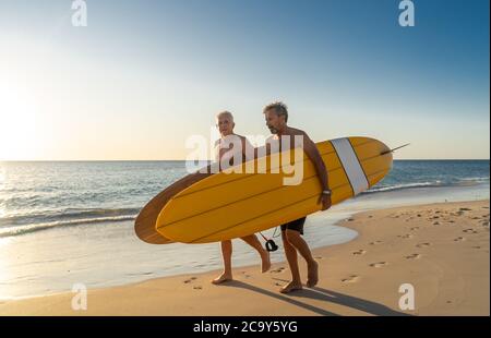 Two mature men walking with surfboards on beautiful beach enjoying paradise and retirement lifestyle. Attractive fit senior adults friends having fun