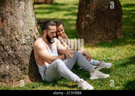 Two sportive heterosexual friends with Tablet PC relaxing on the summer day park lawn after intensive training. Woman with long brown hair wears earbu Stock Photo