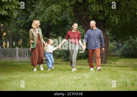 Full length portrait of modern carefree family with two kids holding hands while walking towards camera on green grass outdoors, copy space Stock Photo