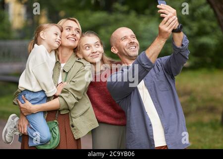 Waist up portrait of modern happy family with two daughters taking selfie photo outdoors while enjoying walk in green park Stock Photo