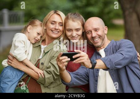 Waist up portrait of modern happy family with two daughters taking selfie outdoors via smartphone while enjoying walk in green park Stock Photo