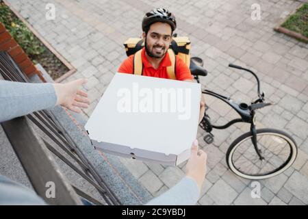 Online order and pizza at home. Smiling courier in helmet, gives boxes to client, near house Stock Photo