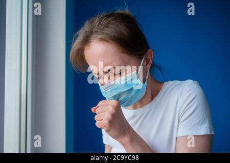 Woman wearing face mask, feeling sick, suffering from coughing, looking out of window in room with blue wall. Self isolation, prevention, quarantine Stock Photo