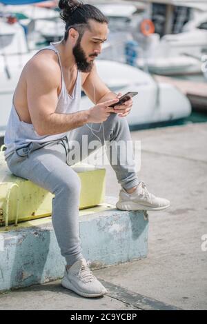 Portrait of sporty healthy strong fitness trainer with beard and hair bun sitting on pier, chatting on smartphone after crossfit outdoor training, wit Stock Photo