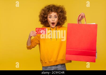 Portrait of shocked surprised curly-haired hipster girl in hoodie holding shopping bags with blank area for advertising text and showing Sale inscript Stock Photo