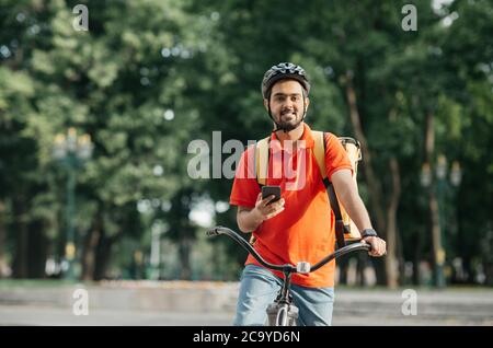 Friendly courier at park, in helmet with backpack on bicycle, holding smartphone and looking for client address Stock Photo