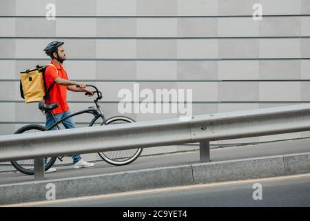 People, delivery and city concept. Courier with helmet and backpack walks along track with bicycle Stock Photo