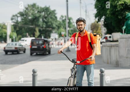 Goods delivery service. Friendly courier in helmet with big bag, stands with bike at crossroads Stock Photo