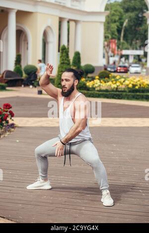 Caucasian athetic man performing squats exercise training in summer park with building on background Stock Photo