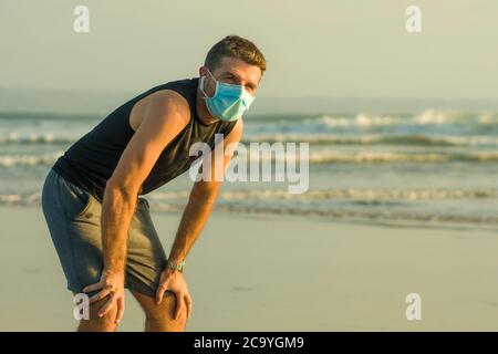 New normal beach jogging workout - young fit and attractive man in face mask breathing tired doing cool off after running on sea training again after Stock Photo