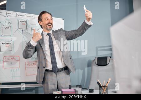 Cheerful man giving thumbs up and making selfie at work Stock Photo