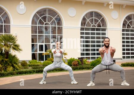 Fitness male and female trainers doing outdoor workout performing lateral lunges and squats in city park, populasing fitness culture among people Stock Photo