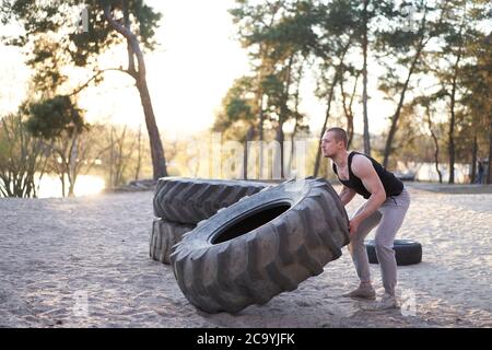 Strong man training workout lifting large tire outdoor DIY gym. Sportive young adult caucasian guy flip big wheel on nature. Handmade equipment sport Stock Photo