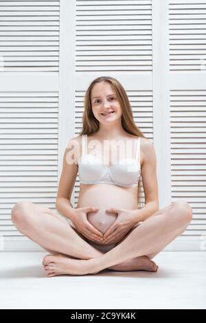 Beautiful young pregnant woman sitting on floor in Lotus position and smiling. Vertical frame. Stock Photo