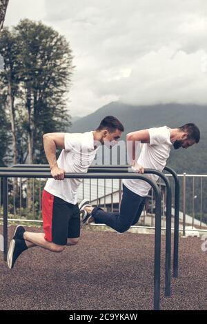 Two crossfit male models in white sportswear doing dips on horisontal bars, warming-up sets before lifting heavy weights. Active and Health Life Conce Stock Photo