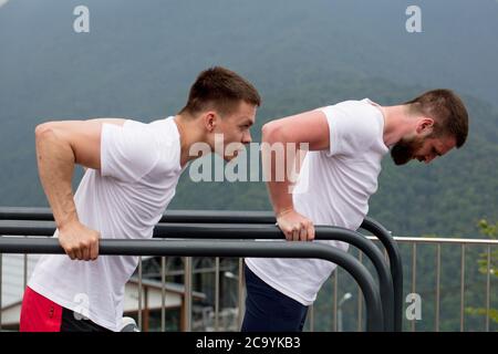 Two crossfit male models in white sportswear doing dips on horisontal bars, warming-up sets before lifting heavy weights. Active and Health Life Conce Stock Photo