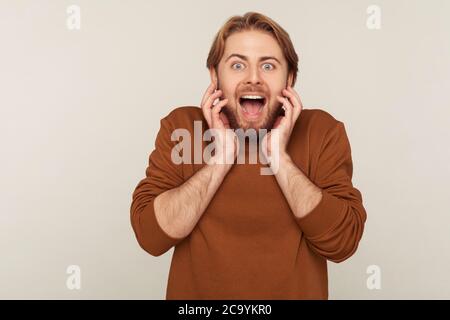 Portrait of surprised bearded man standing with open mouth and screaming in sudden shock, touching face in amazement, astonished by unbelievable succe Stock Photo