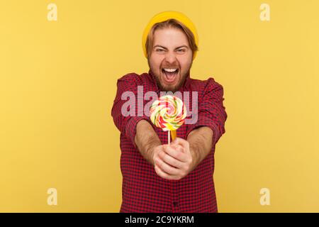 Extremely joyful delighted hipster guy in beanie hat, checkered shirt giving lollipop to camera and screaming with excitement, crazy about sweet sugar Stock Photo