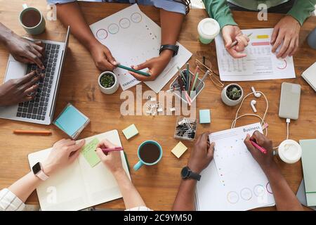 Top view close up of multi-ethnic group of people working together at cluttered wooden table with coffee cups, mugs and stationary items, teamworking or studying concept, copy space Stock Photo