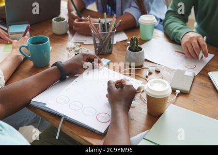 Close up of multi-ethnic group of people working together at cluttered table with cups, mugs and stationary items, teamworking or studying concept, copy space